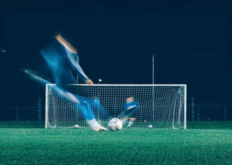 Fußballspieler schießt bei Nacht mit Bewegungsunschärfe einen Ball ins Tor auf einem beleuchteten Platz.